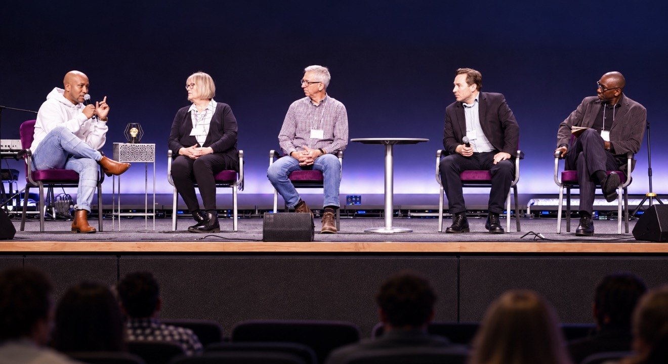 Andrew Alleyne in a white hoodie speaking with a microphone during a five-person panel conversation on stage