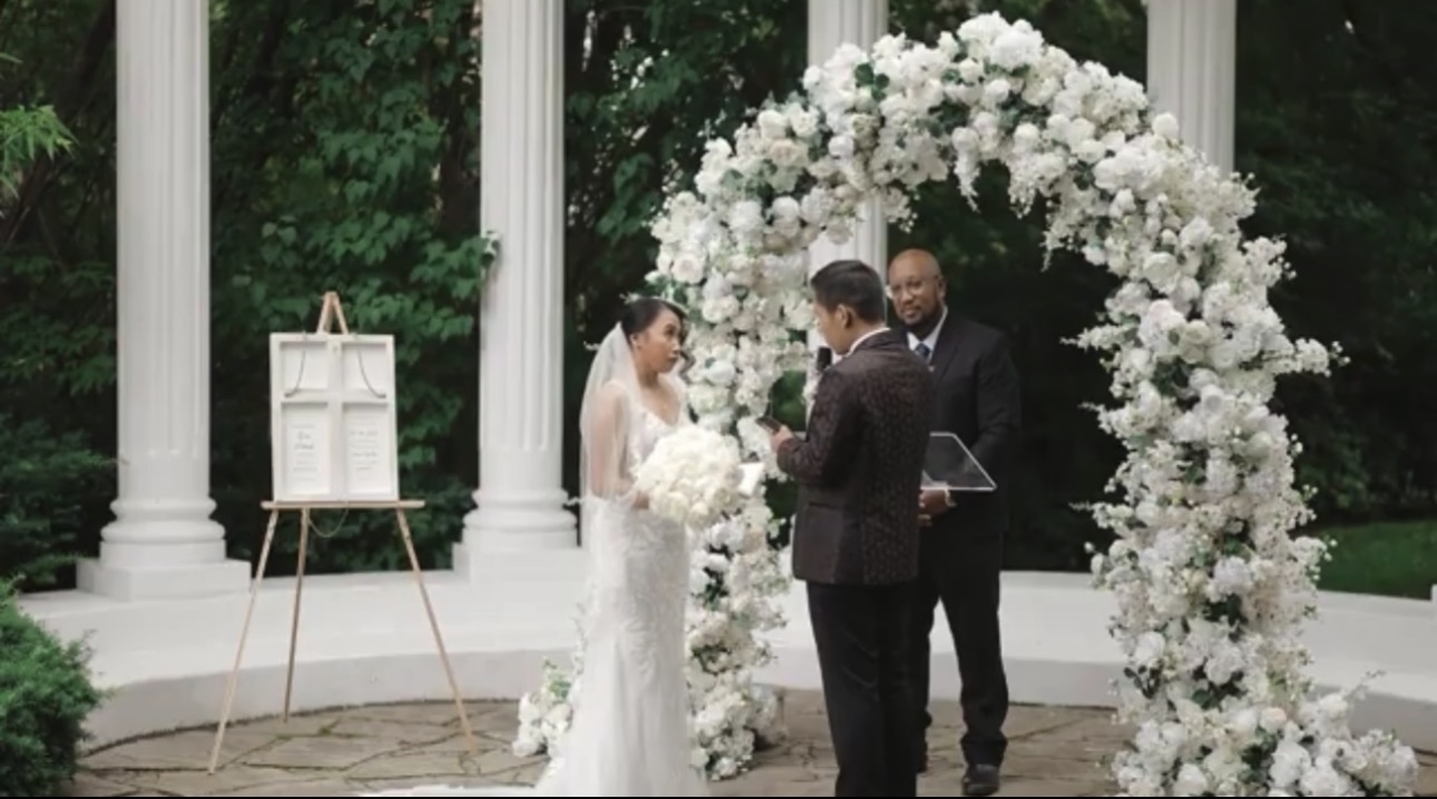 Andrew Alleyne officiating an outdoor ceremony beneath a white floral arch