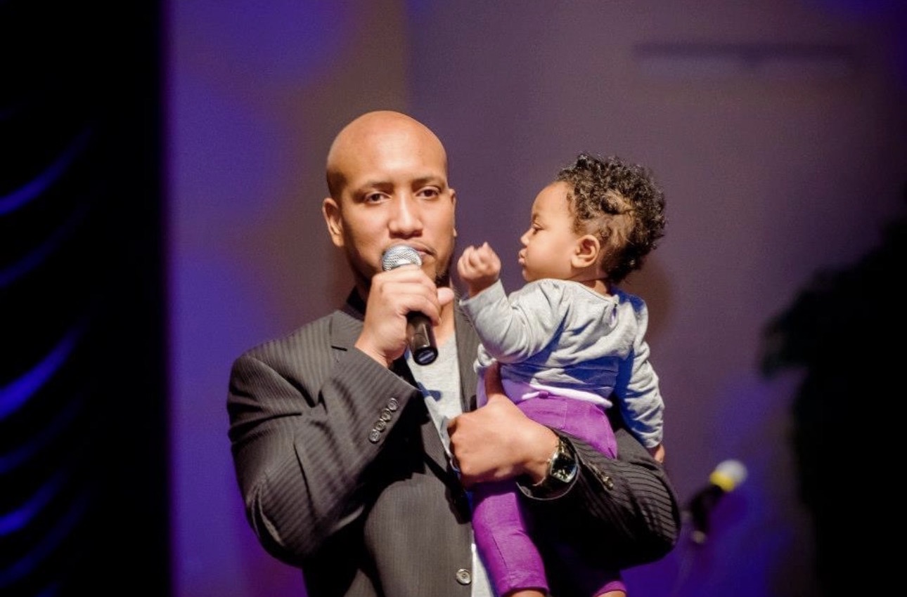 Andrew Alleyne speaking on stage while holding one of his children