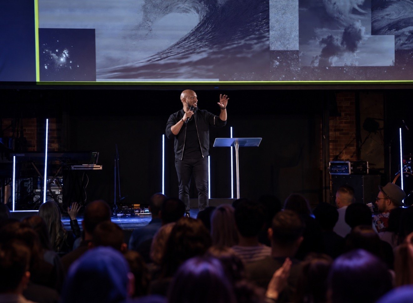 Andrew Alleyne preaching on a large stage with one hand raised beside a clear podium, a congregation gathered in the foreground