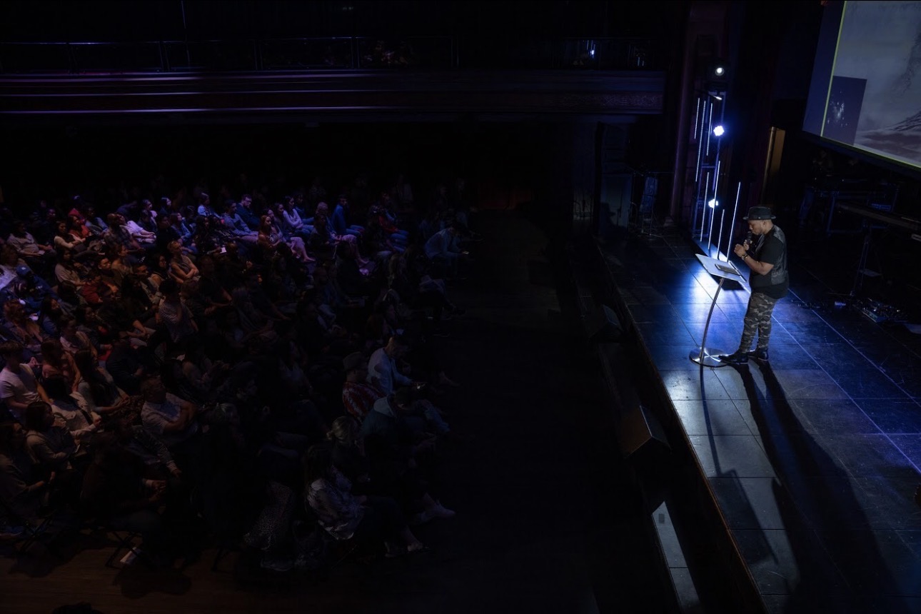 Andrew Alleyne preaching from a lit lectern to a darkened theatre full of people