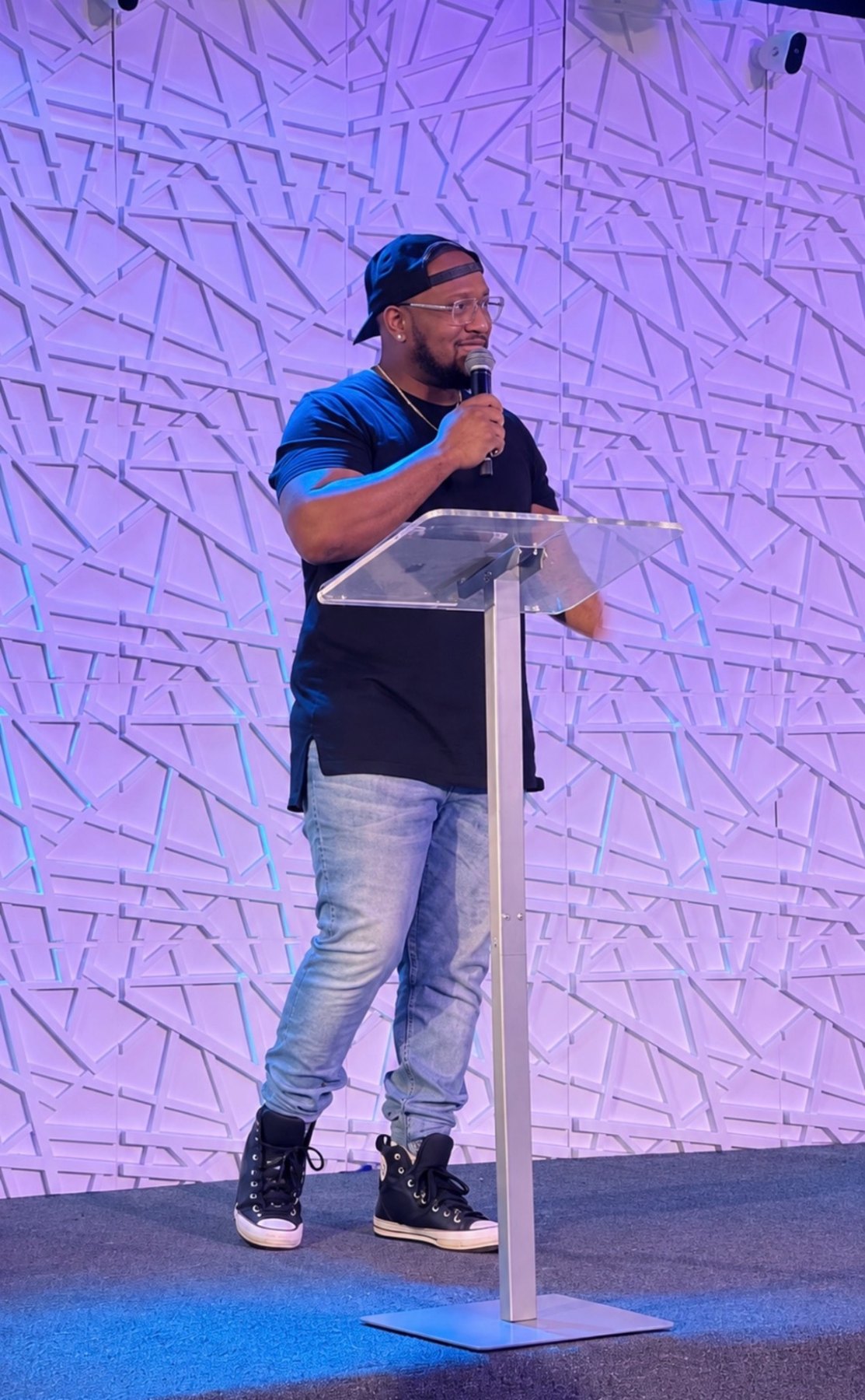 Andrew Alleyne in a backwards cap and glasses speaking with a microphone behind a clear podium against a purple-lit geometric wall